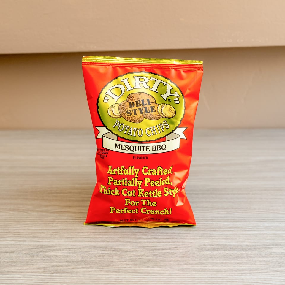 A red bag of Dirty Deli Style Mesquite BBQ Potato Chips stands upright on a light wooden surface.