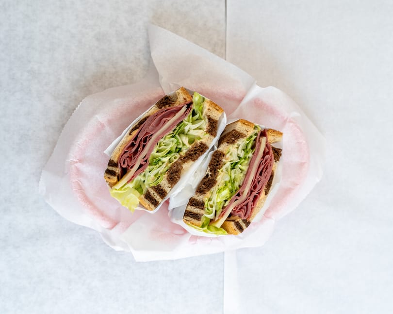 Two deli sandwiches with meat, cheese, and shredded lettuce on marbled rye bread, wrapped in paper.