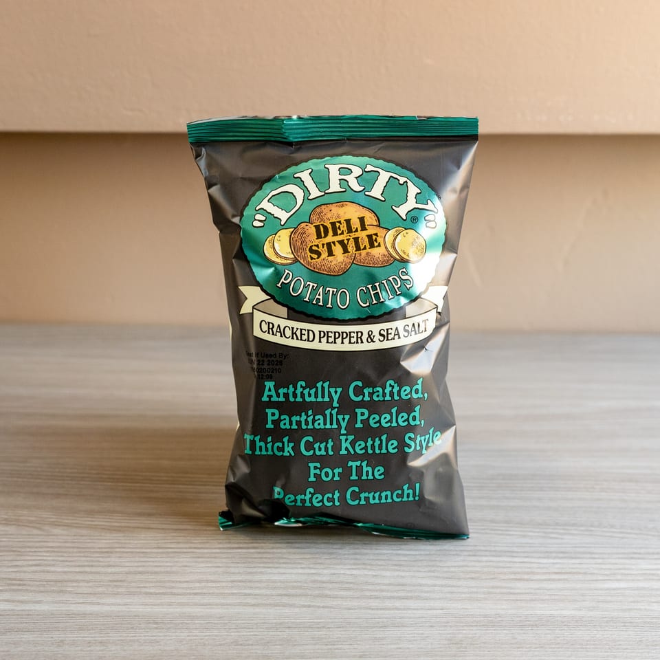 A bag of Dirty Deli Style Potato Chips, Cracked Pepper & Sea Salt flavor, on a wooden surface.