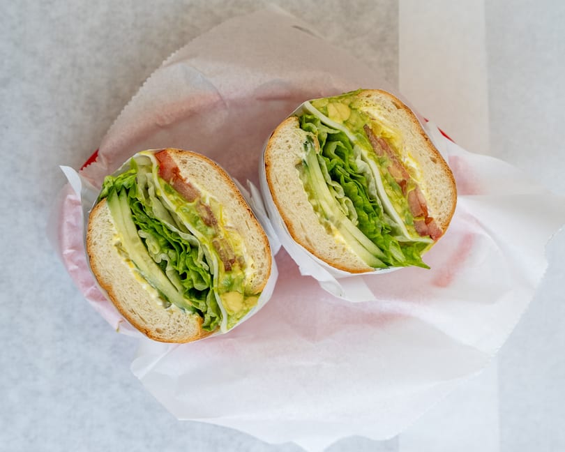 Two halves of a sandwich filled with lettuce, cucumber, tomato, and avocado, wrapped in paper.